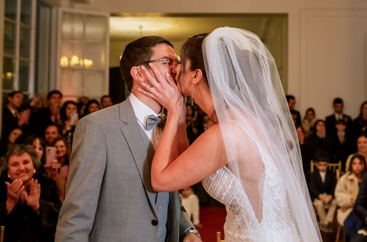 Bride and groom share first kiss at wedding ceremony with guests applauding in elegant venue.