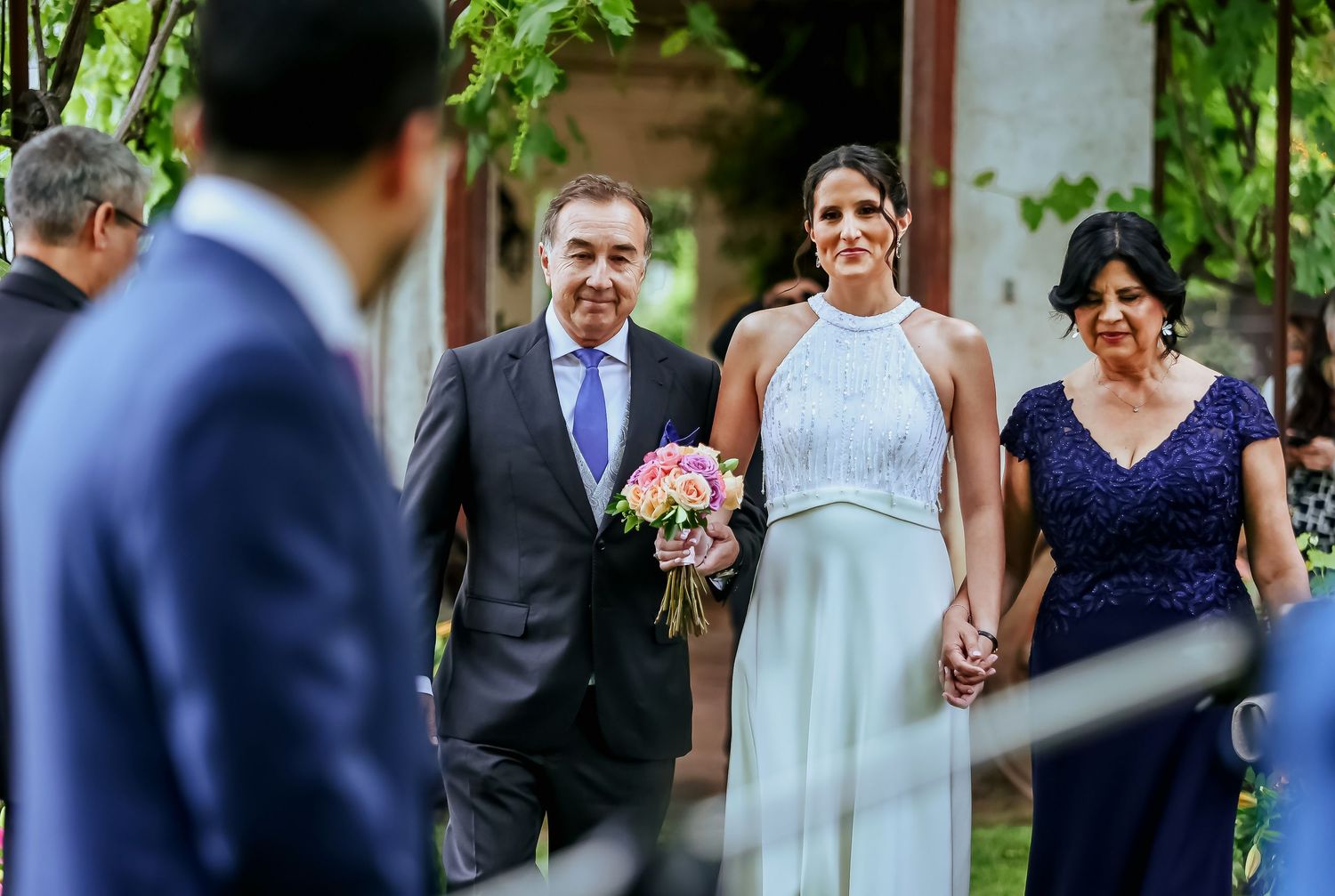 Bride in white halter gown walks down aisle with parents at outdoor garden wedding ceremony.