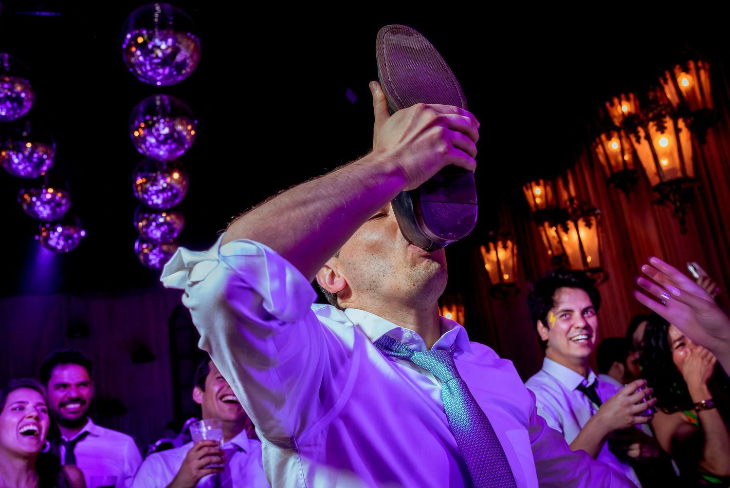 Smiling man drinks from a large shoe while surrounded by laughing guests at a wedding reception.
