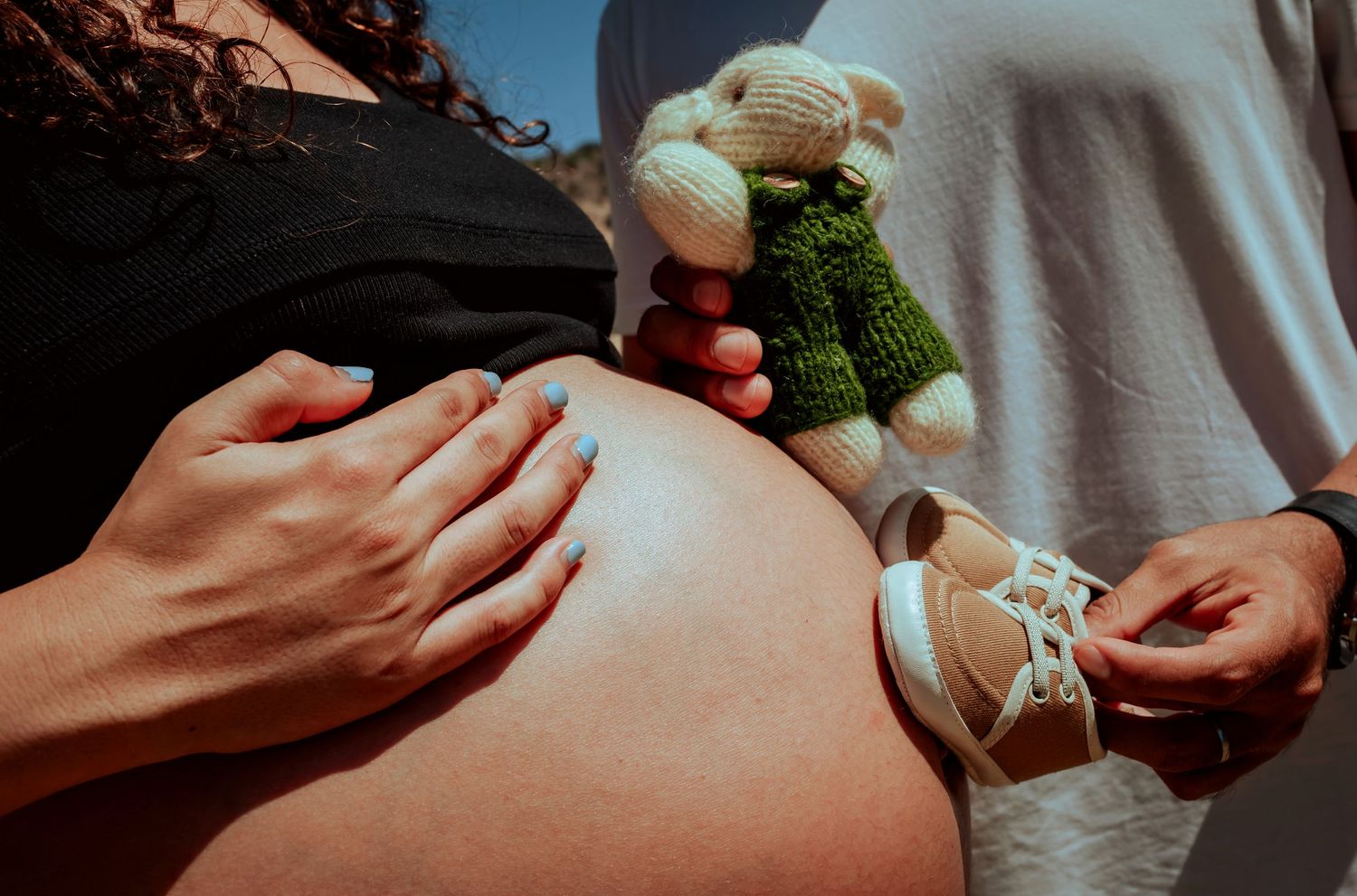 Close-up of pregnant belly with knitted teddy bear and tiny baby shoes held by expecting couple.