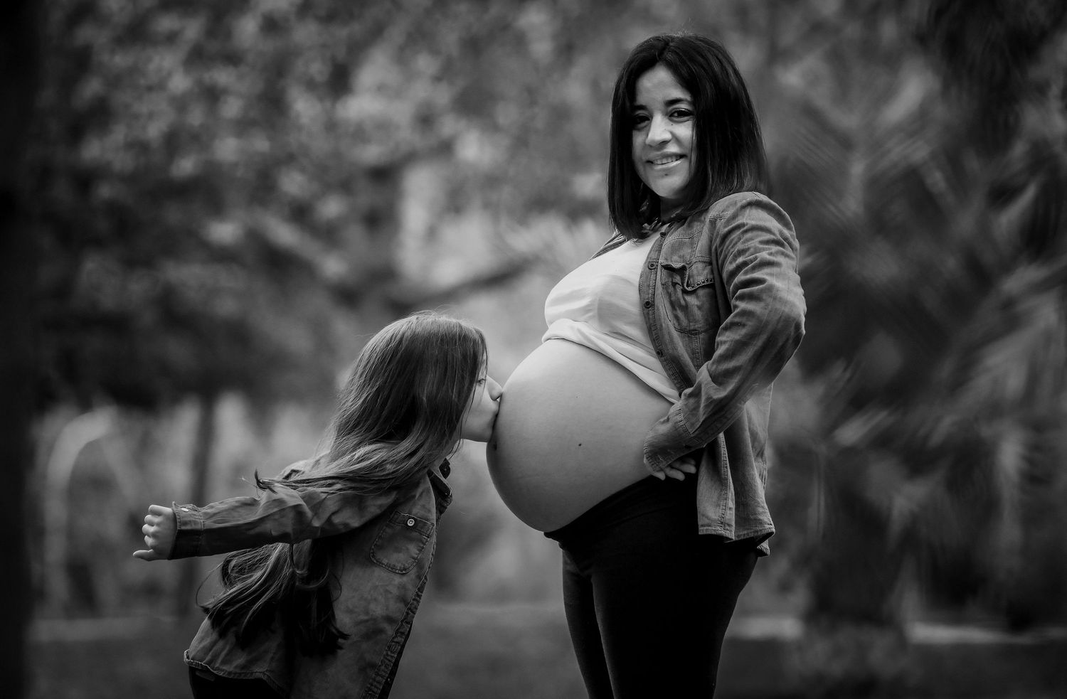 Black and white photo of smiling pregnant woman with child hugging her baby bump in park.