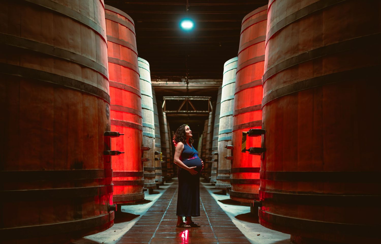 Pregnant woman in blue dress stands between large wooden wine barrels in red-lit cellar.