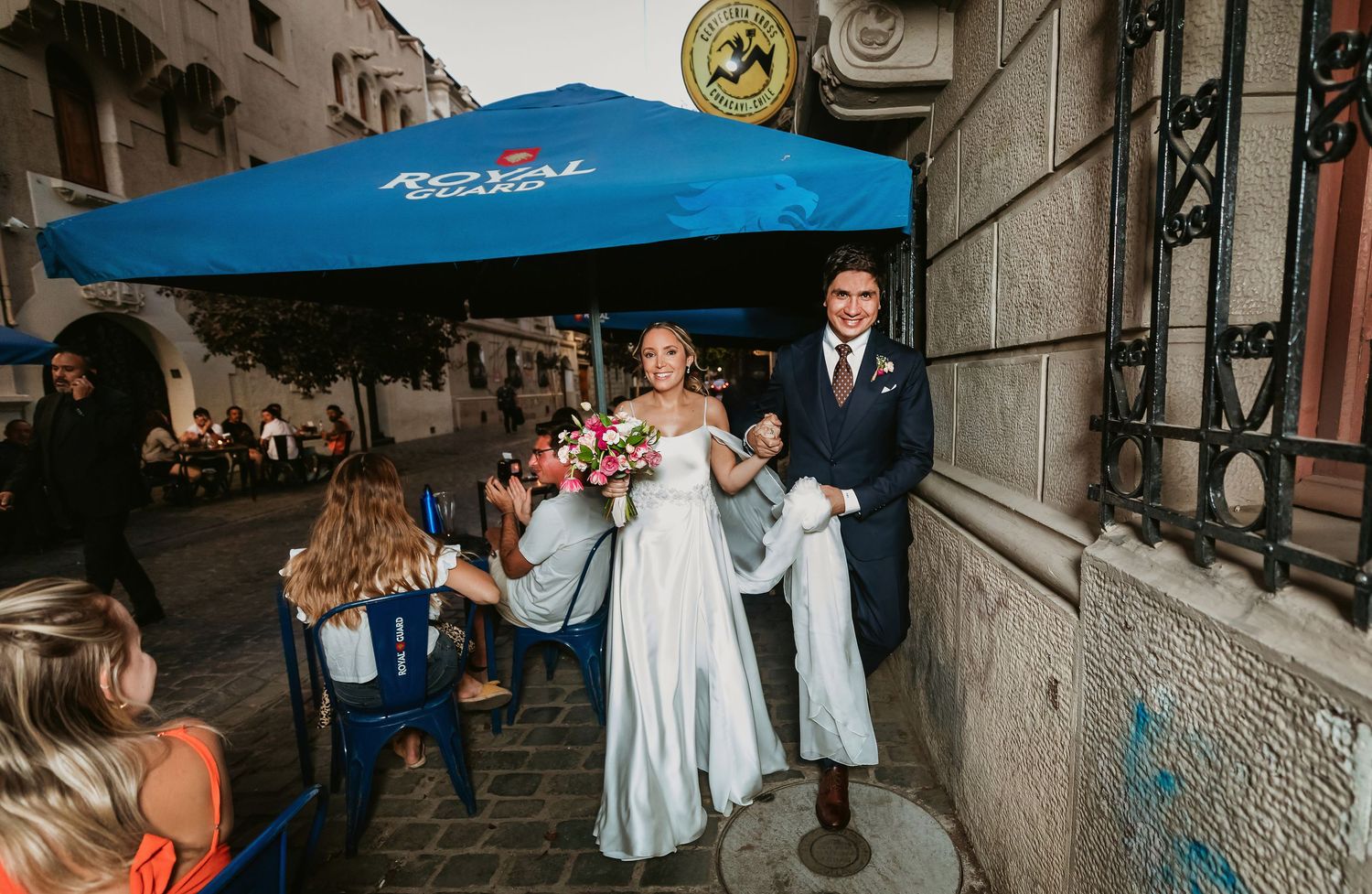 Smiling newlyweds walking past outdoor cafe under blue Royal Studio umbrella on city street.