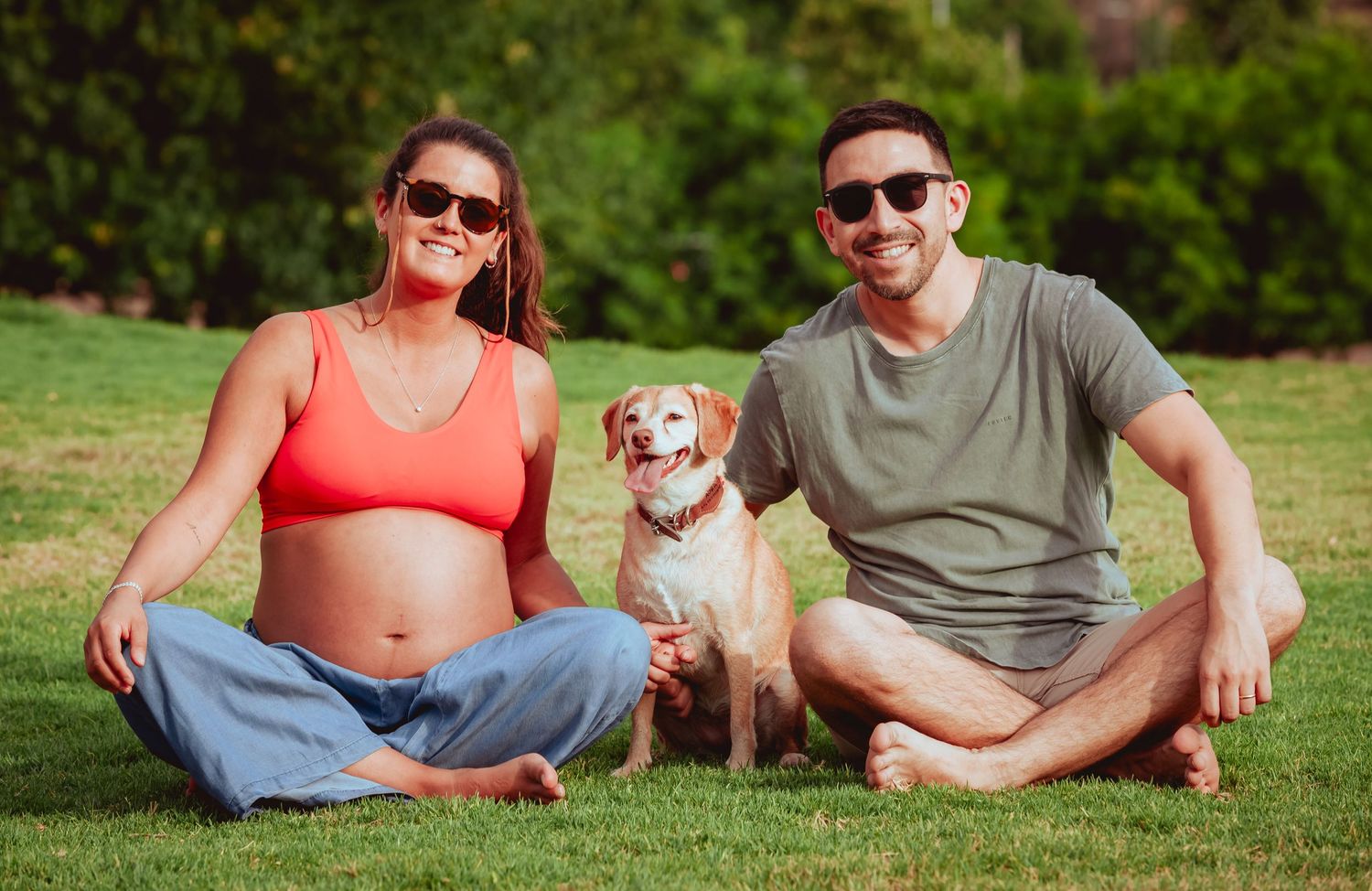 Smiling expectant couple sitting on green grass with their golden Beagle dog between them on sunny day.