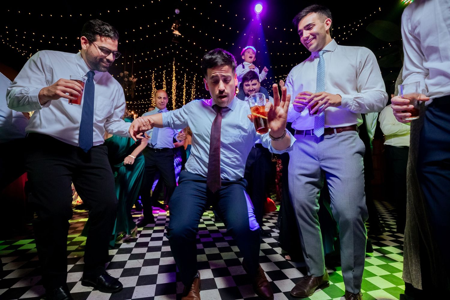 Groomsmen energetically dance together on checkered dance floor at lively wedding reception party.