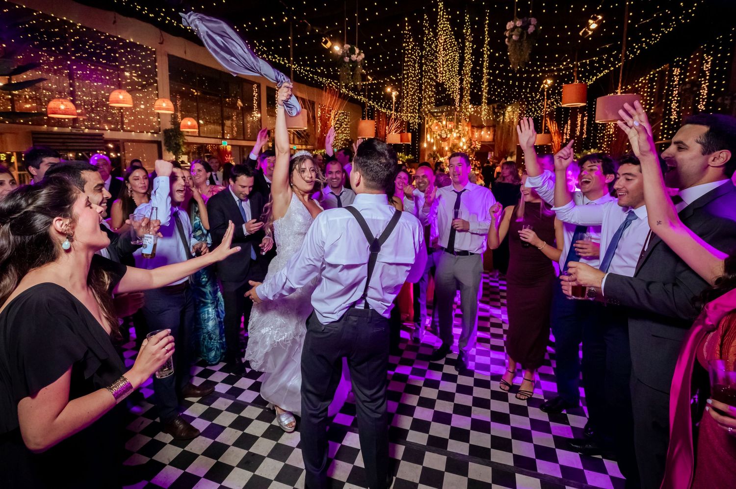 Bride and groom share a kiss on a checkered dance floor surrounded by celebrating wedding guests.