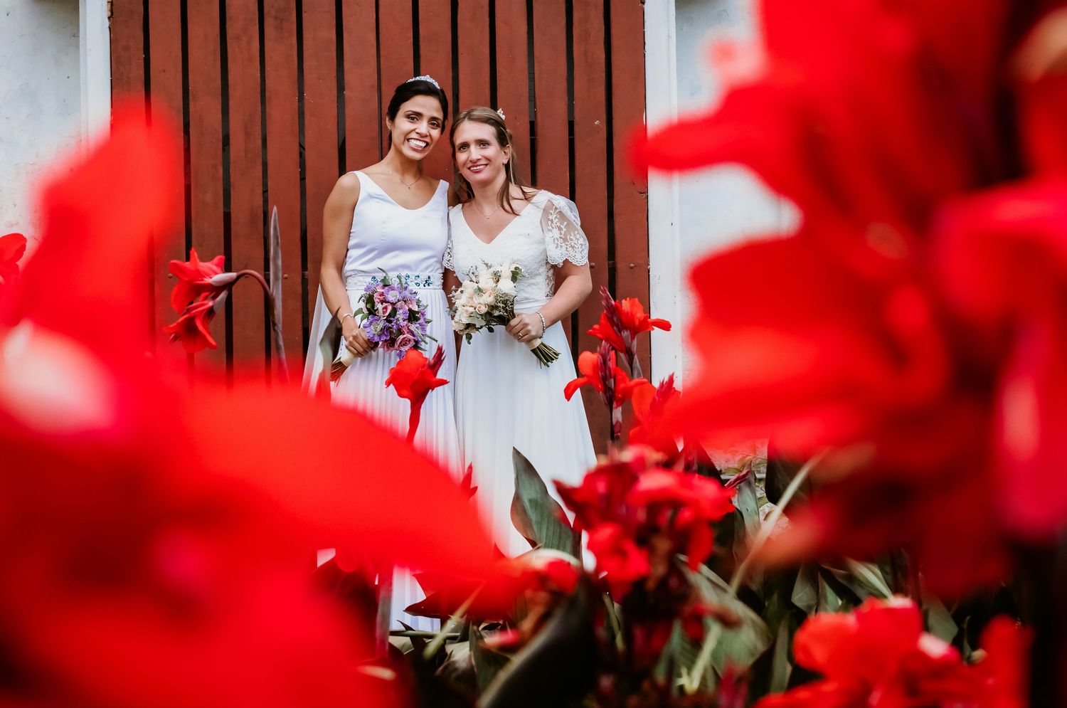Two smiling brides with bouquets standing by red wooden door framed by bright red flowers.