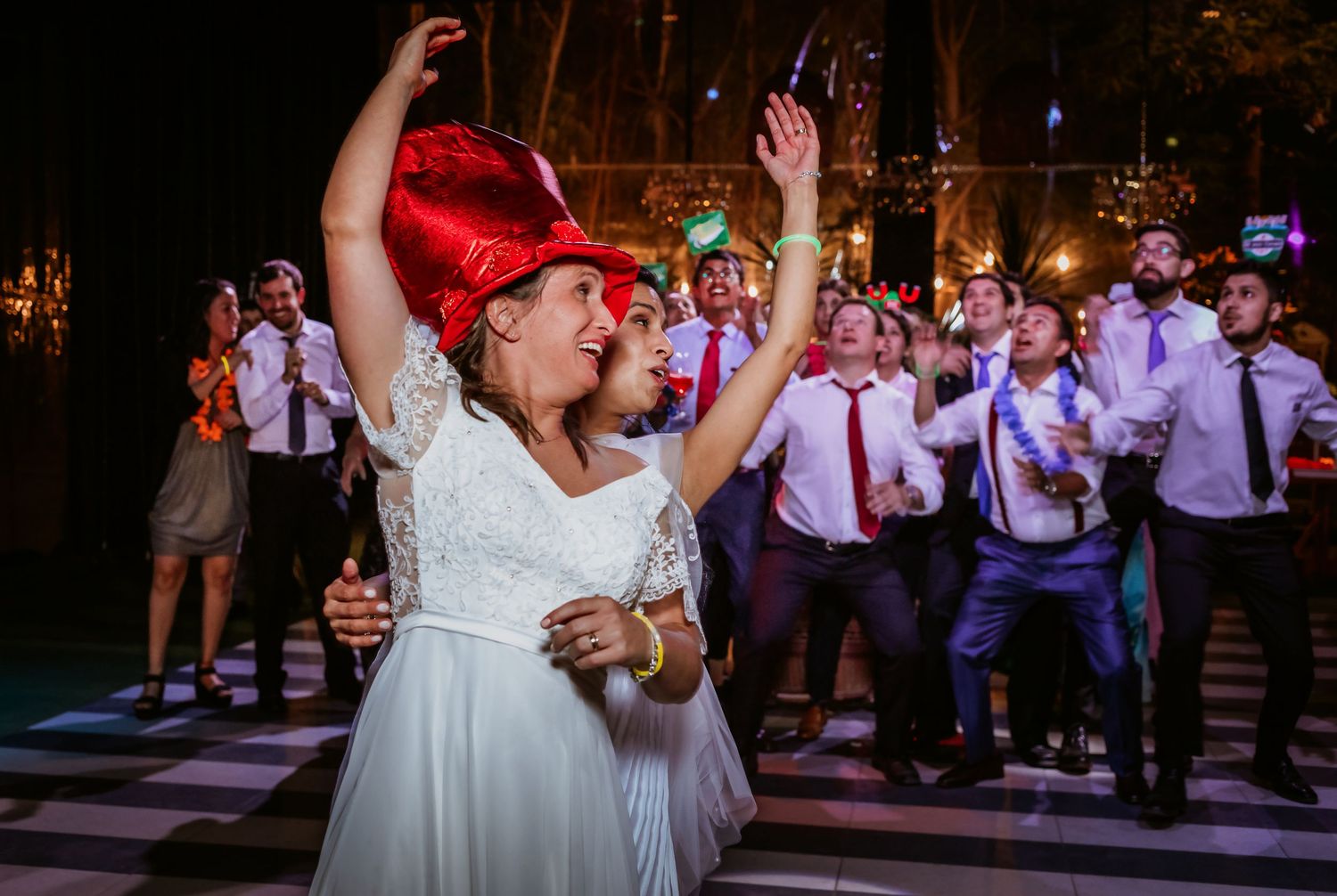 Bride in white gown and red hat celebrates with arms raised among cheering wedding guests on dance floor.