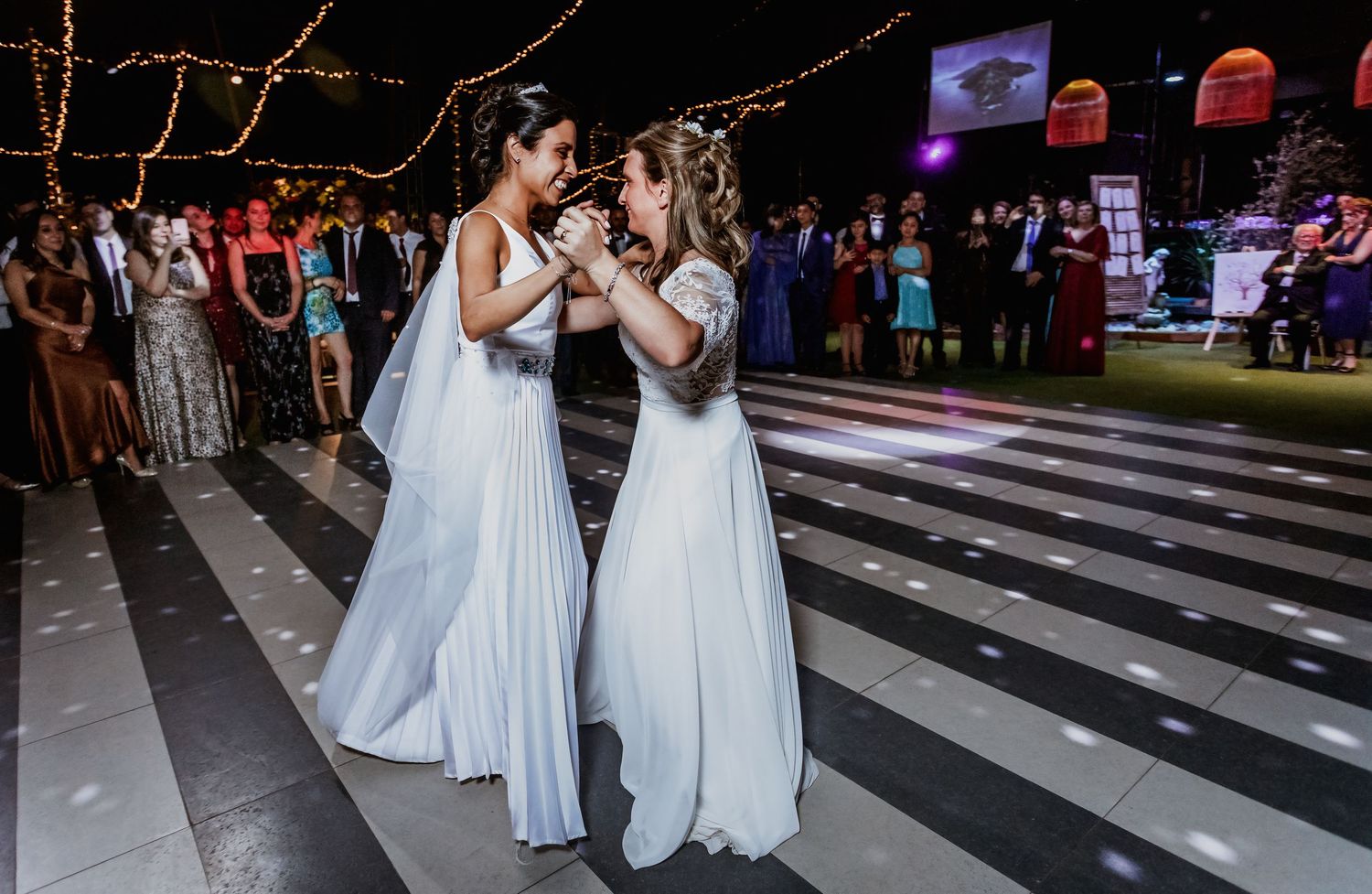 Two brides hold hands during emotional first dance on striped wedding reception dance floor.