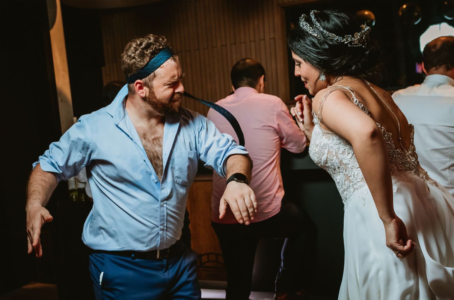 Bearded man in blue shirt dancing with bride in white gown at wedding reception.