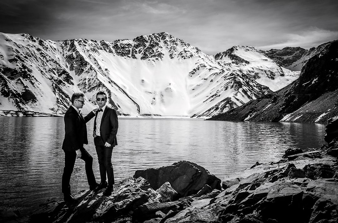 Black and white photo of two men in formal suits standing by a dramatic glacial mountain lake.