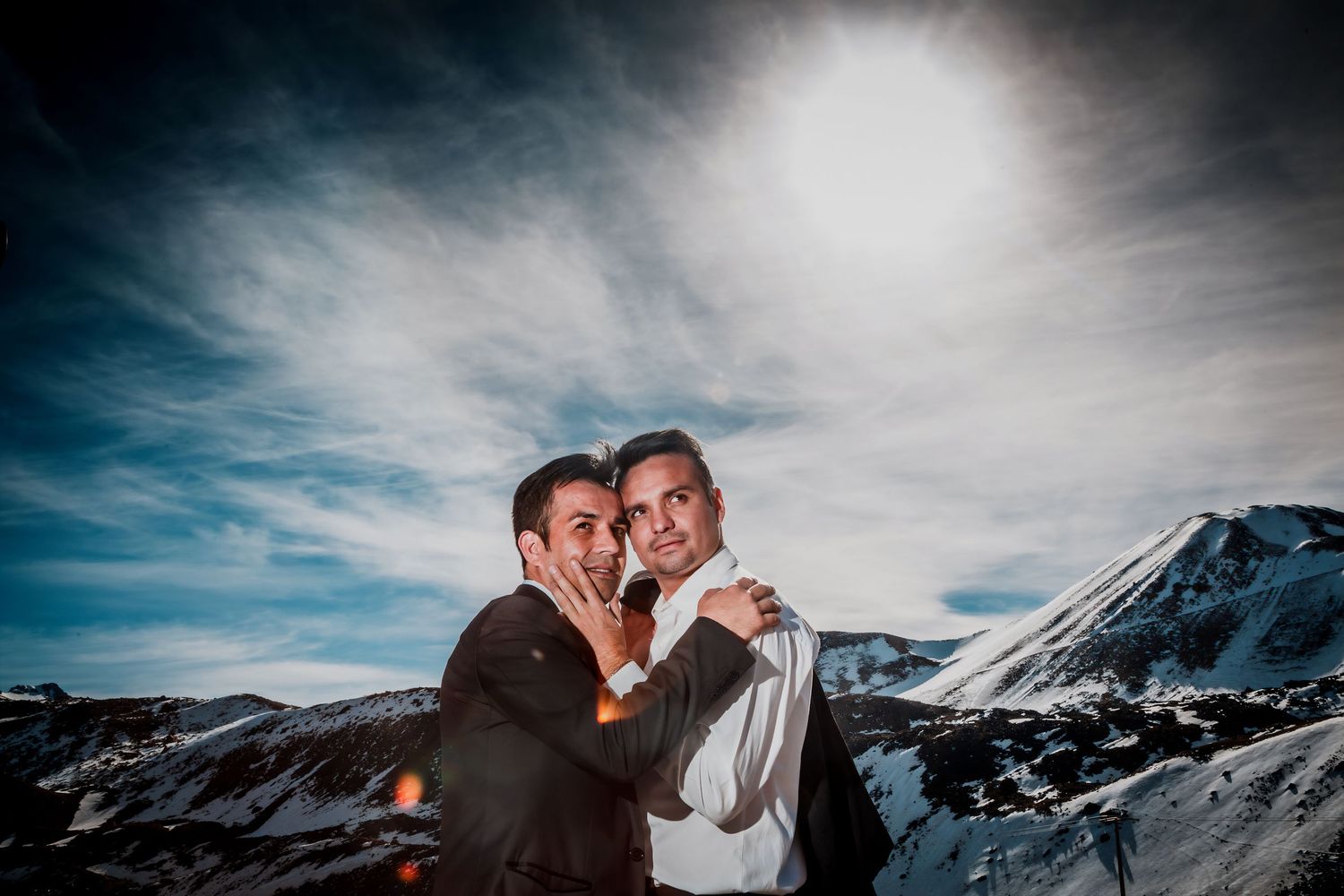 Two grooms share a joyful romantic moment with snow-capped mountains and dramatic sky in the background.