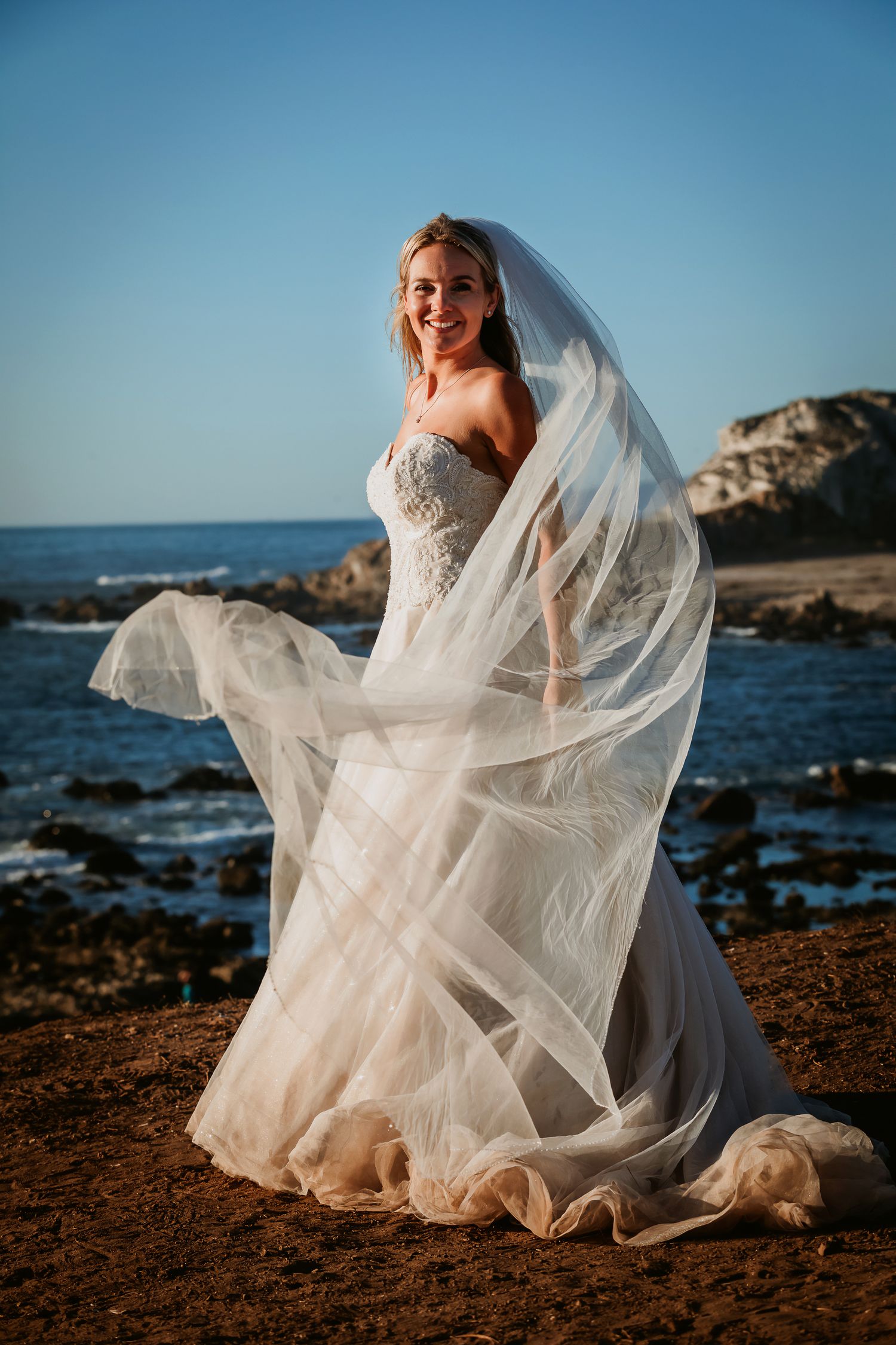 Joyful blonde bride in lace strapless gown with veil flowing in wind on scenic coastal shoreline.