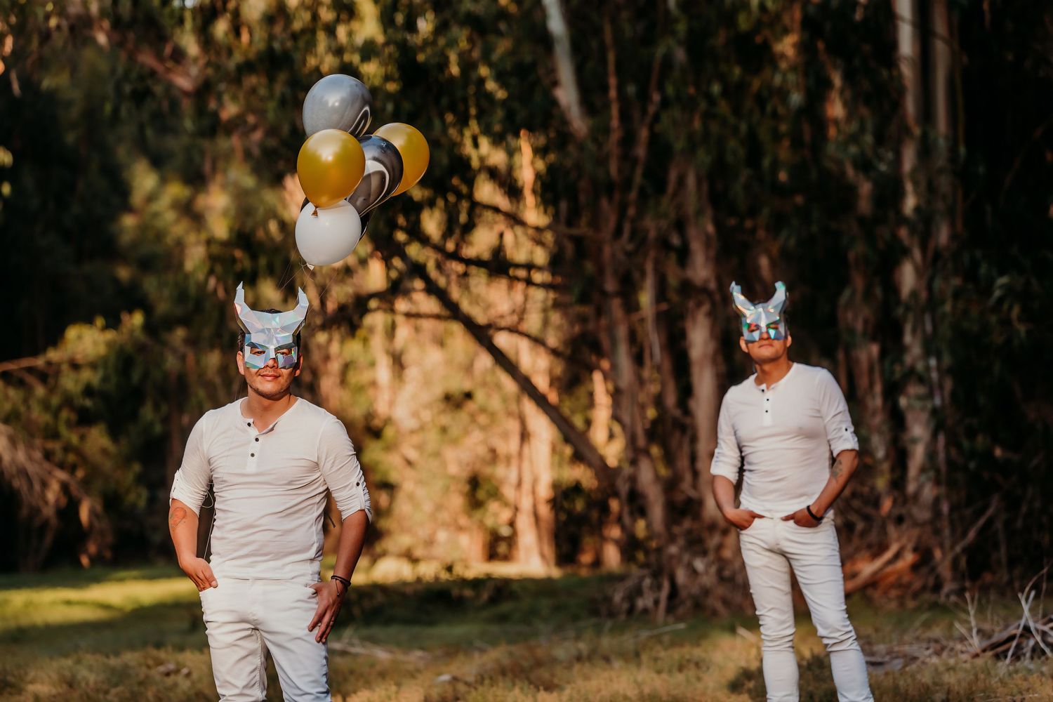 Two men wearing Viking masks and white clothes stand outdoors holding colorful balloons near trees.