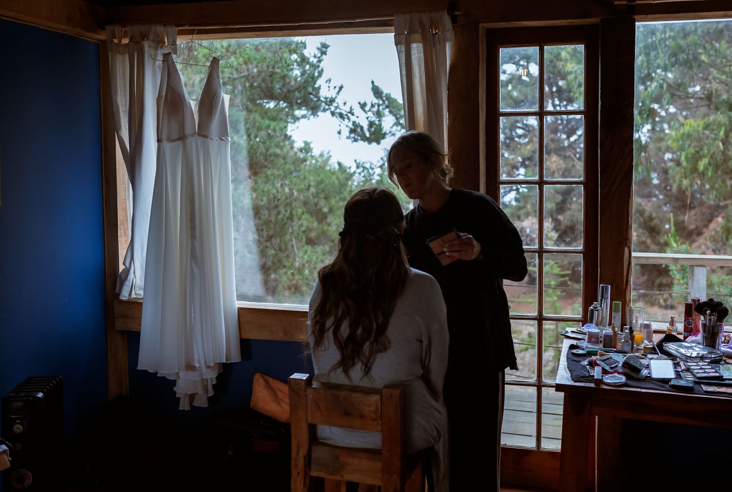 Bride sits as a stylist assists her with hair and makeup, wedding dress hanging by the window.