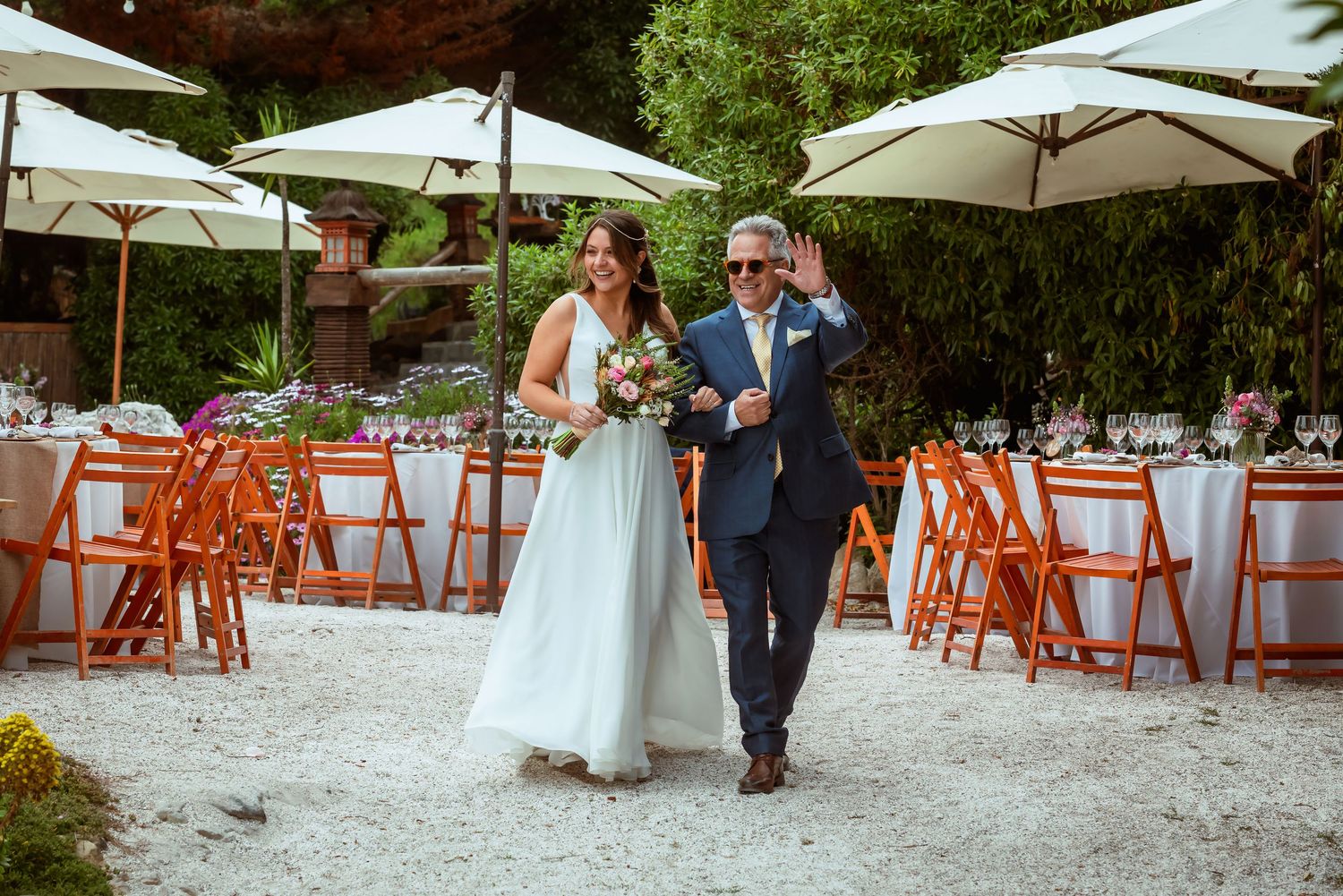 Bride and escort walk through outdoor wedding venue with orange chairs and umbrellas.