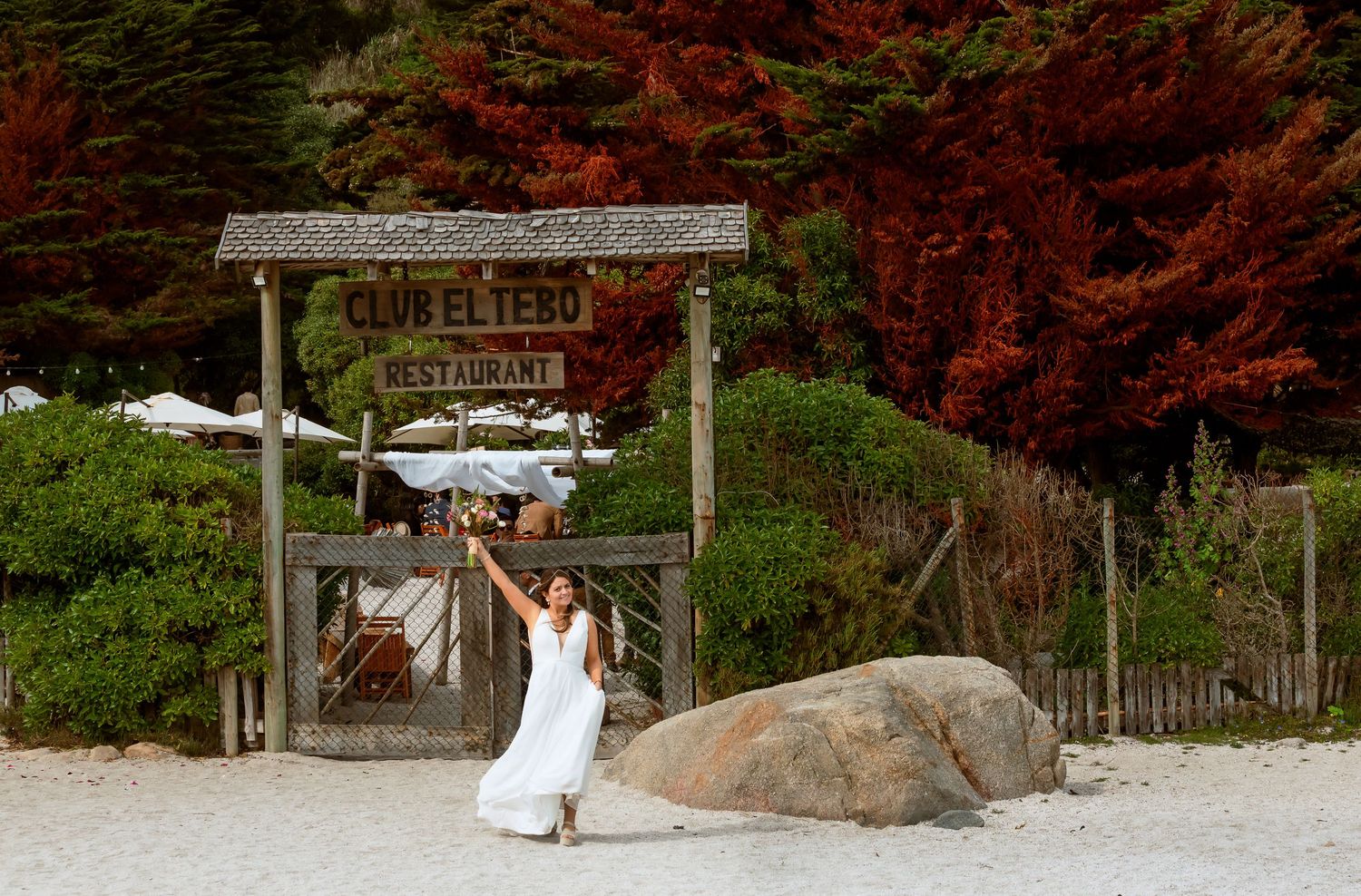 Woman in white dress posing at entrance of Club El Tero Restaurant on a sandy beach surrounded by trees.