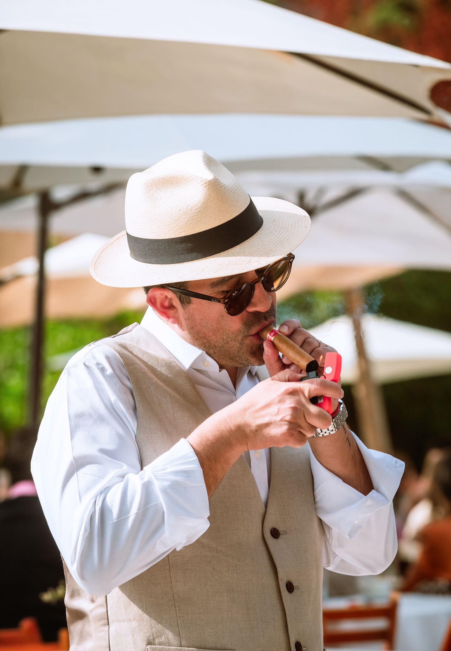 Man in panama hat and sunglasses lighting a cigar at an outdoor event in bright sunlight.