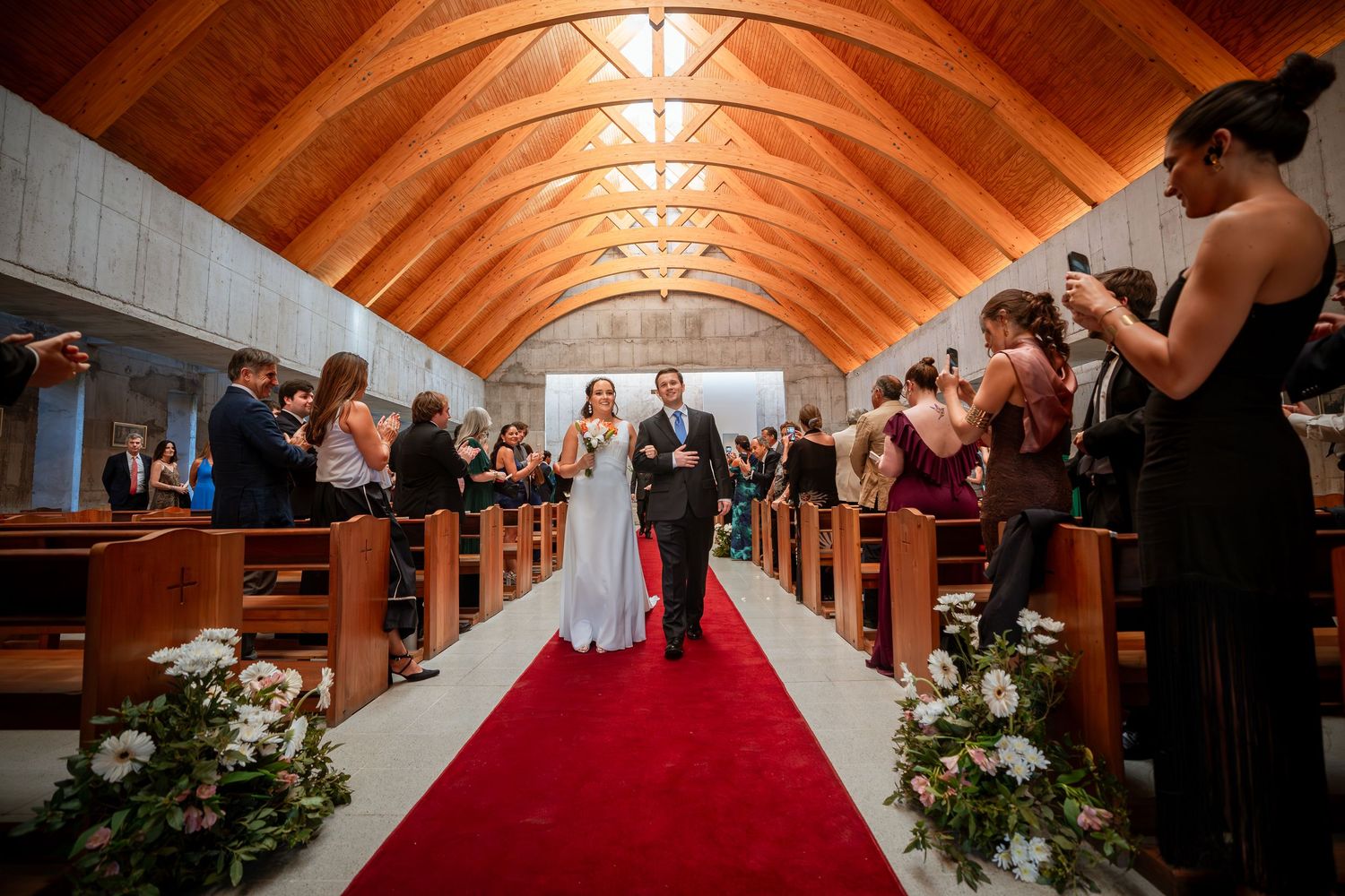 Newlyweds walk down red carpet aisle amid cheering guests in wooden-ceiling church.