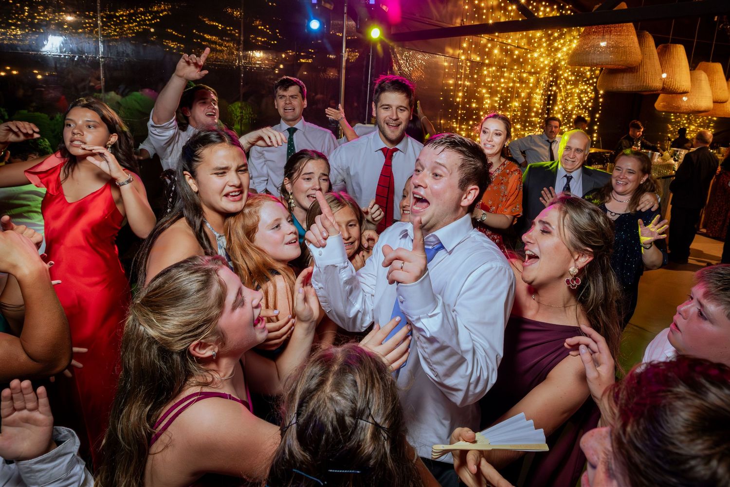 Groom sings surrounded by enthusiastic cheering wedding guests at lively outdoor reception celebration.