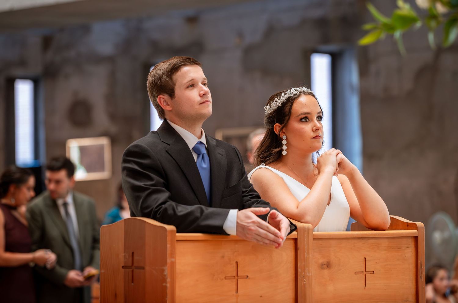 Bride and groom kneel together at wooden pew with crosses during their Catholic wedding ceremony.