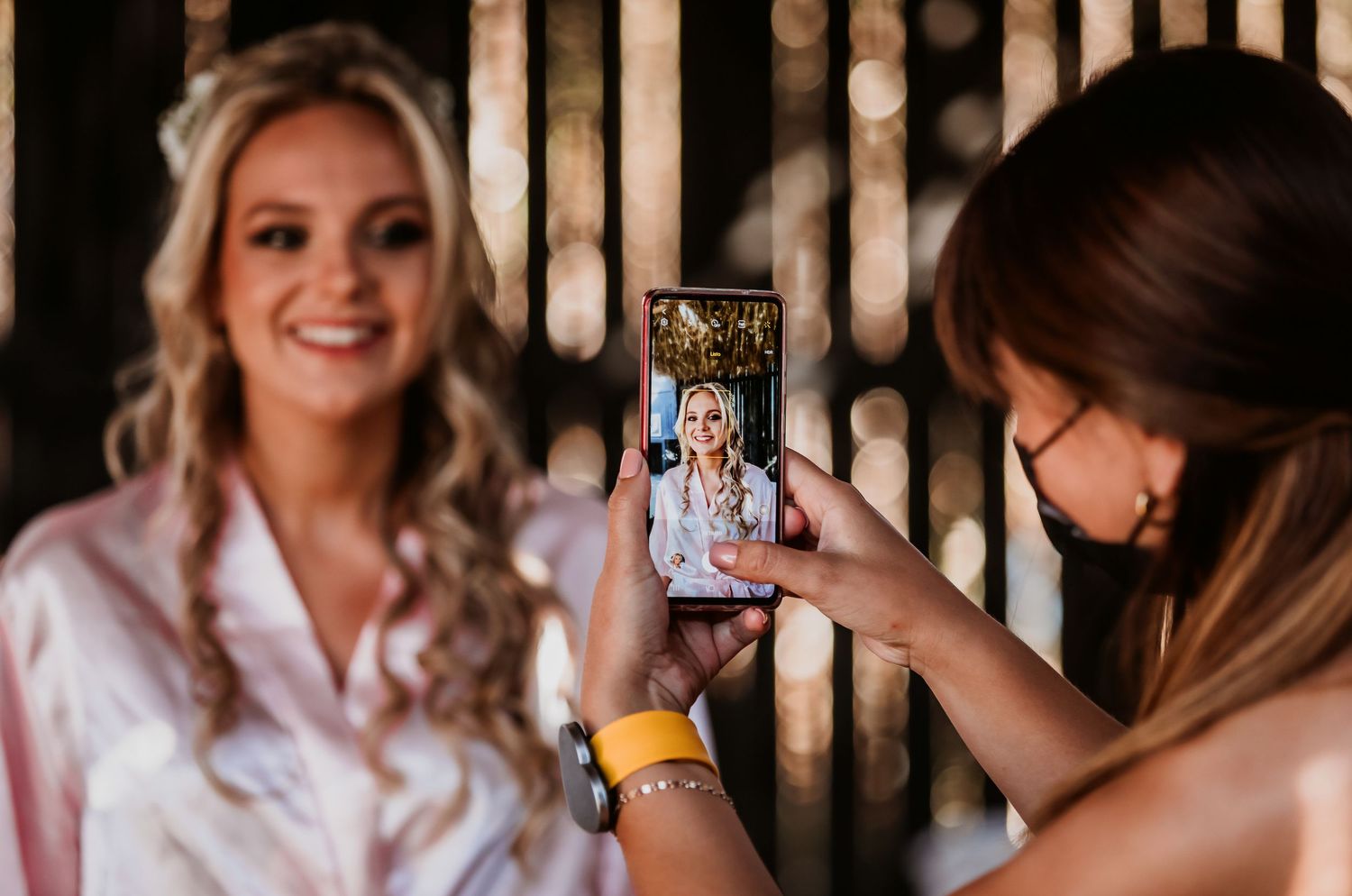 Bridesmaid photographing smiling bride in pink satin robe with phone at wedding preparation.