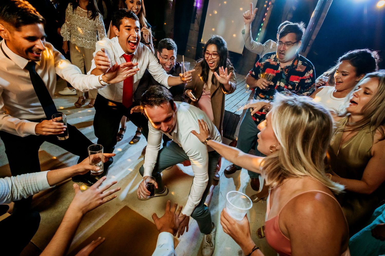 Excited wedding guests surround groom on dance floor during lively reception.