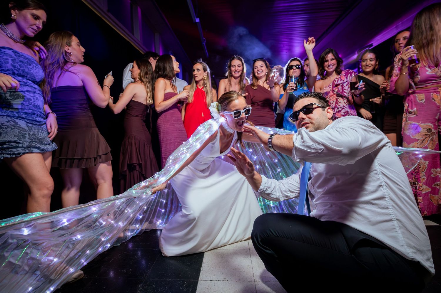 Bride and groom pose dramatically on dance floor with LED wings at lively wedding reception.