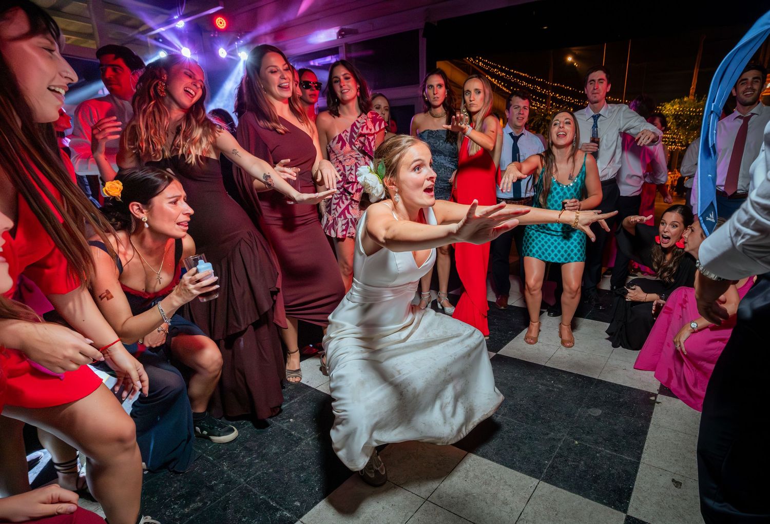 Laughing bride in white gown crouches low on dance floor surrounded by cheering wedding reception guests.