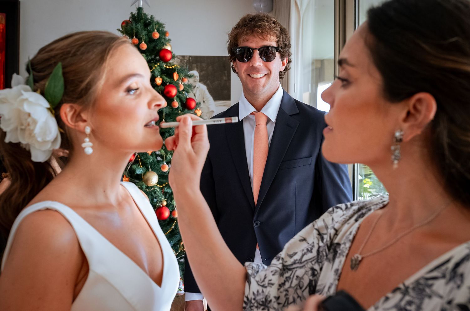 Makeup artist applying lipstick to bride near Christmas tree as man in sunglasses watches.