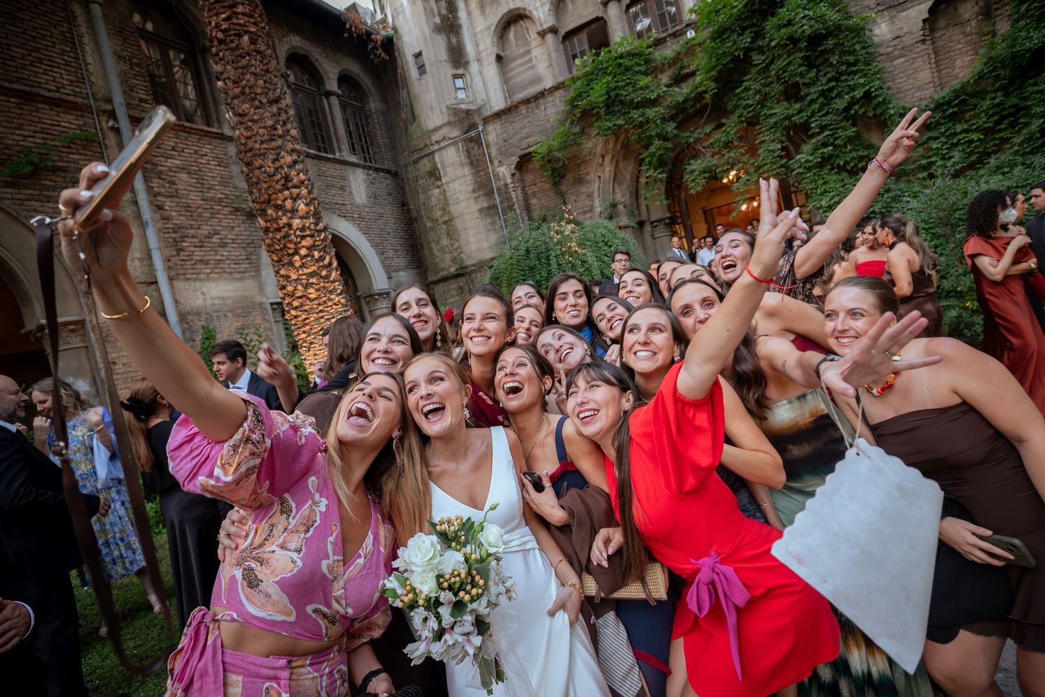 Bride takes joyful group selfie with smiling wedding guests in sunlit outdoor venue.