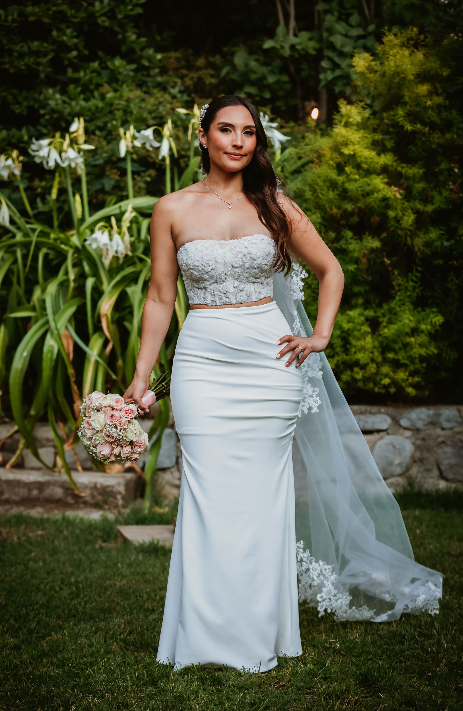 Bride in white two-piece strapless gown with lace top, holding bouquet, standing in garden with white flowers.