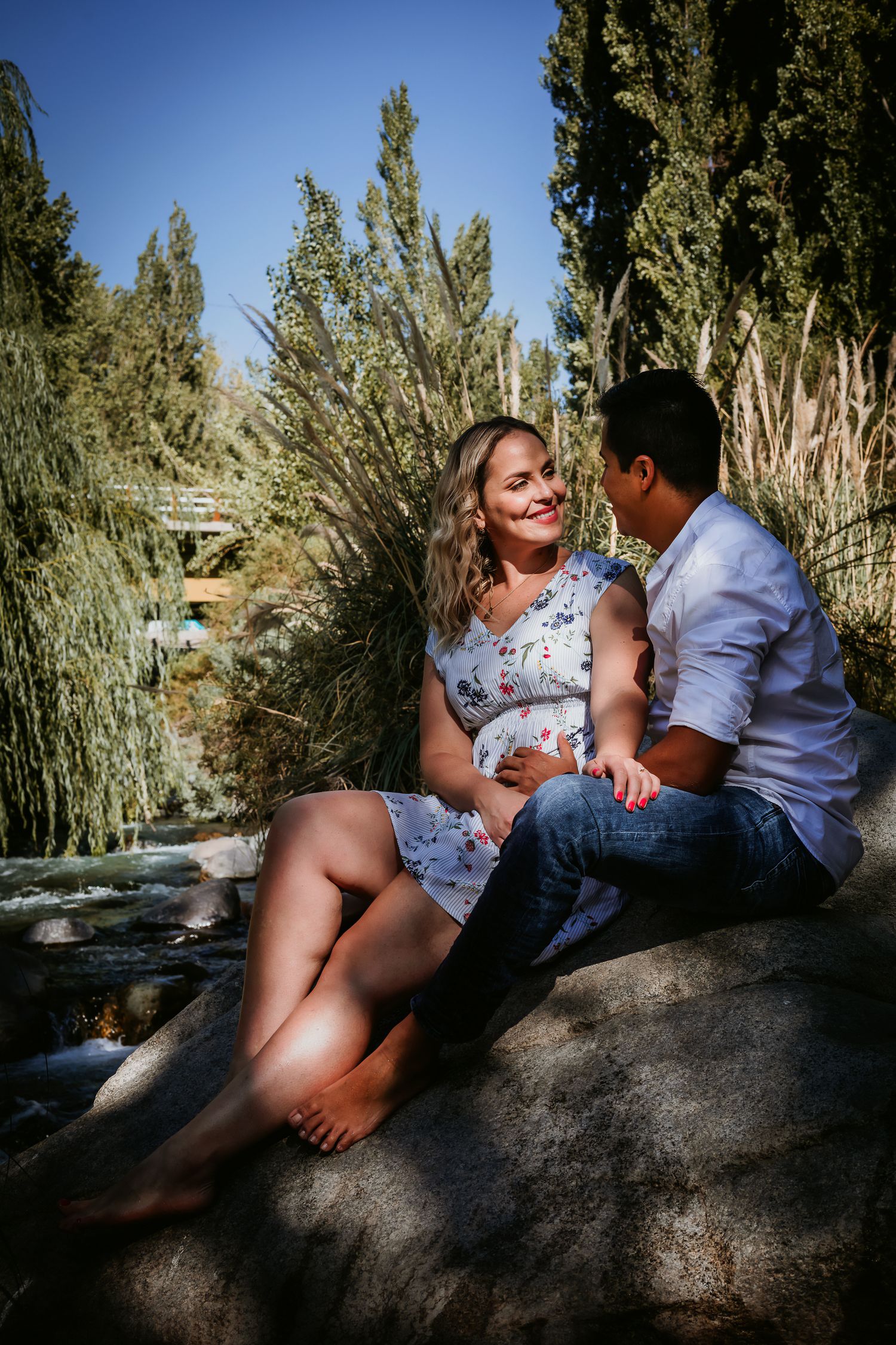 Pregnant couple sitting on rocks in a lush garden, sharing a tender moment outdoors on a sunny day.