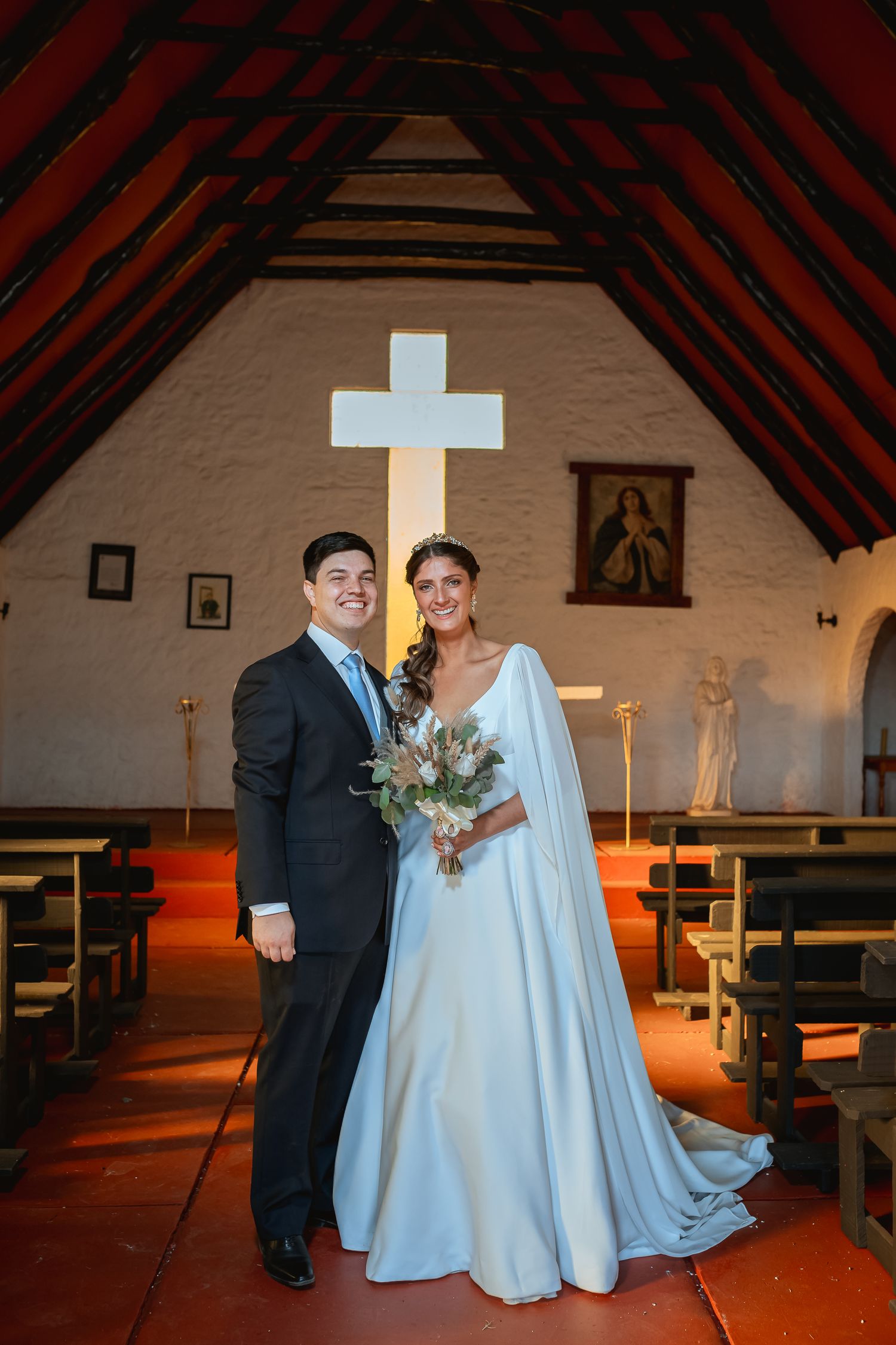 Bride and groom posing together inside a small chapel with a white cross and wooden beamed ceiling on their wedding day.