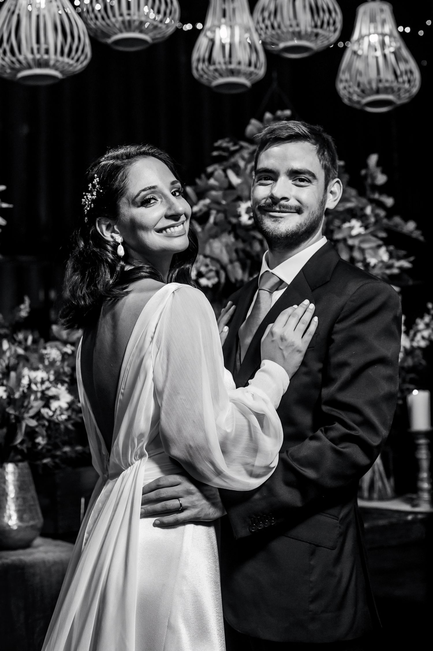 Black and white wedding photo of smiling bride in backless gown and groom in suit embracing at elegant reception.