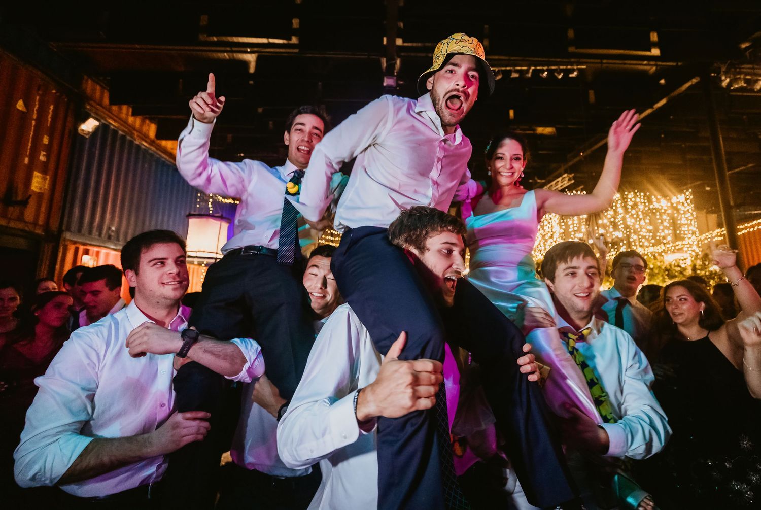 Joyful wedding reception guests celebrating with someone lifted on shoulders on a lively dance floor.