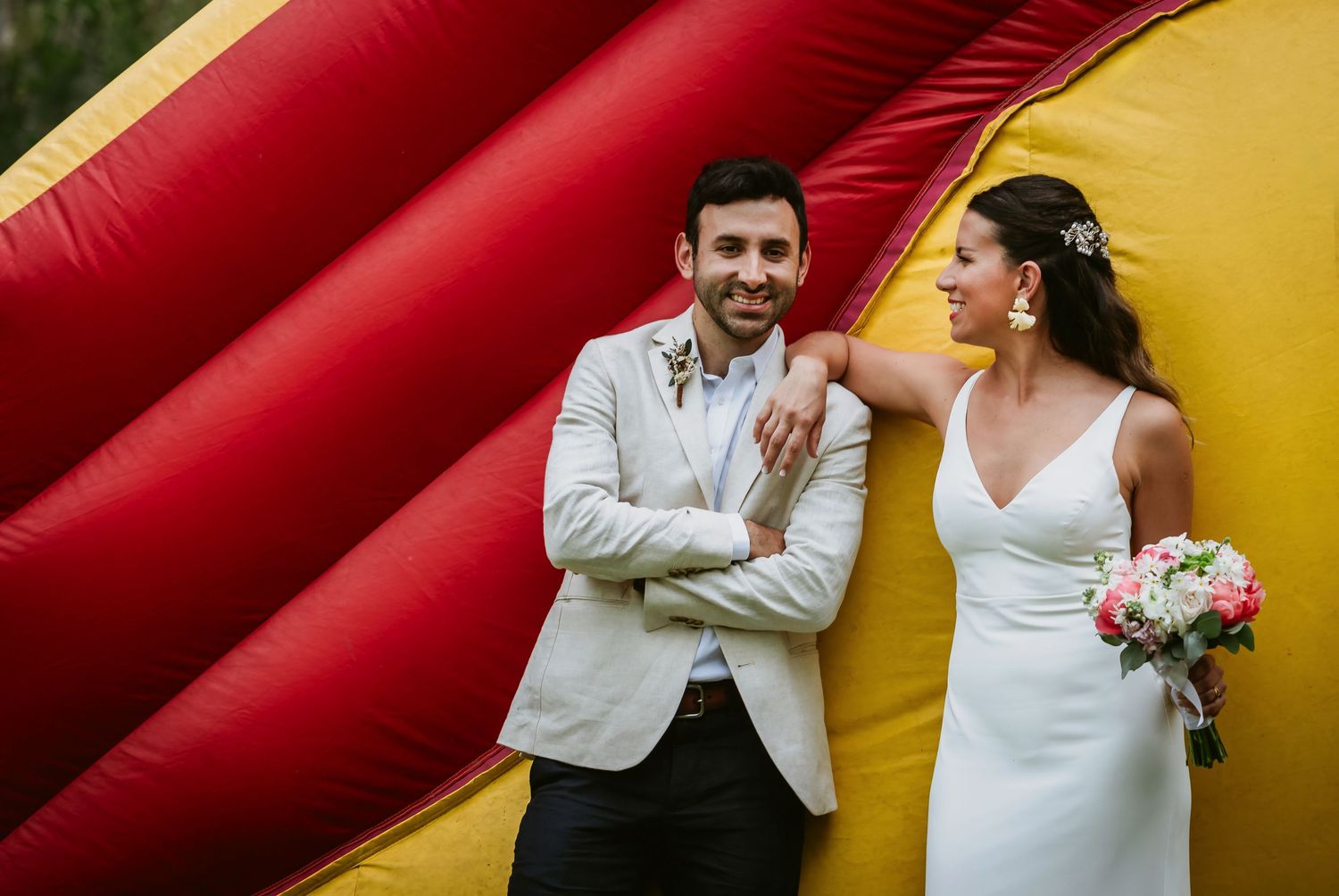 Bride and groom laughing together in front of a colorful red and yellow inflatable bouncy castle at their wedding.