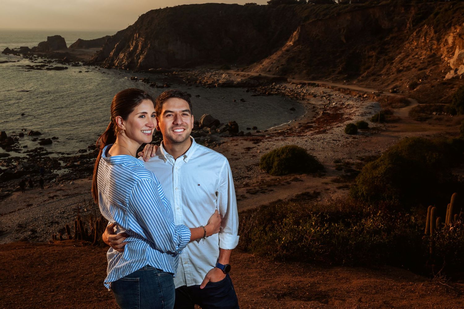Happy couple embracing on coastal cliffside with dramatic rocky shoreline at sunset.