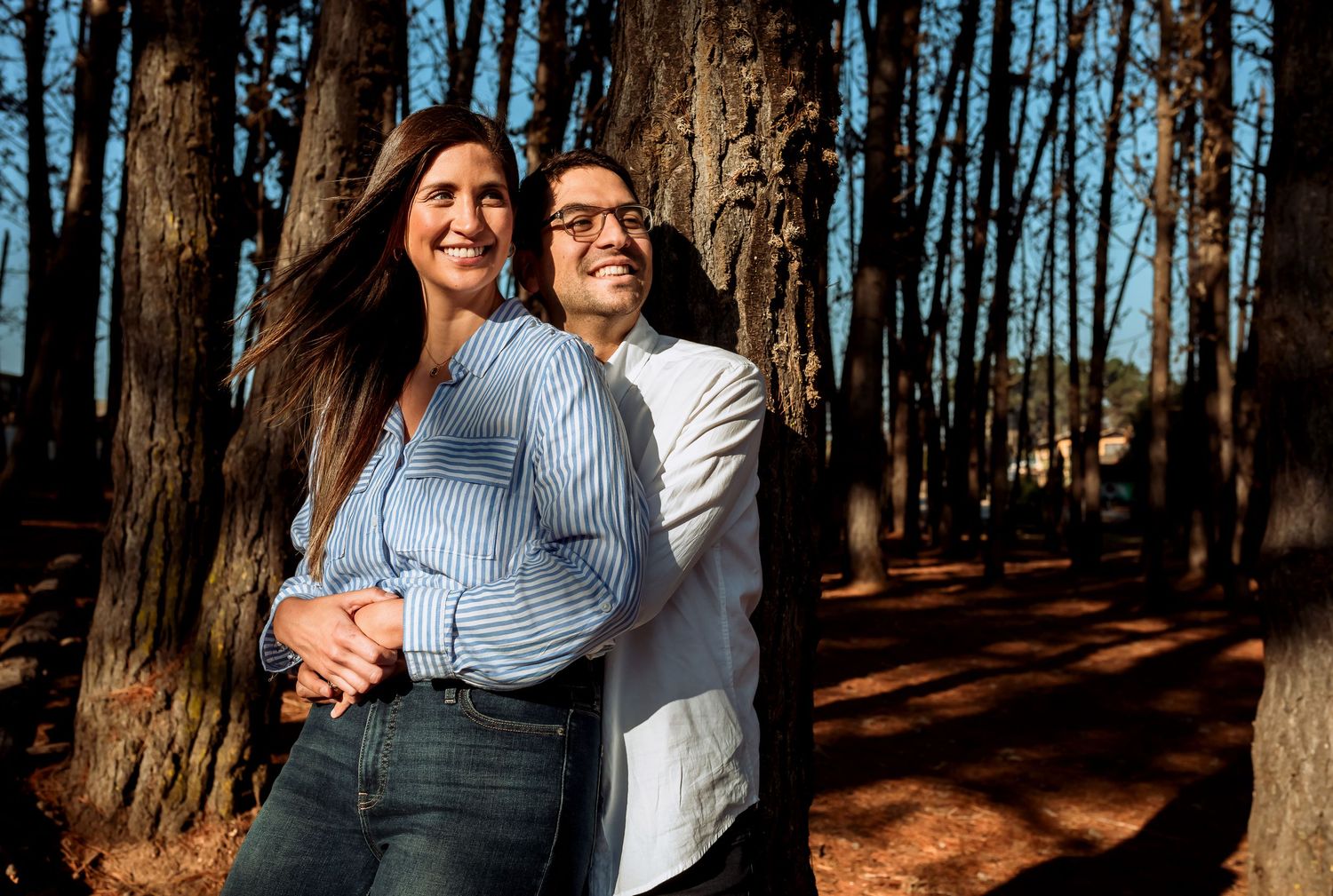 Couple embracing and smiling in sunlit pine forest with tall trees and warm light during outdoor engagement photoshoot.