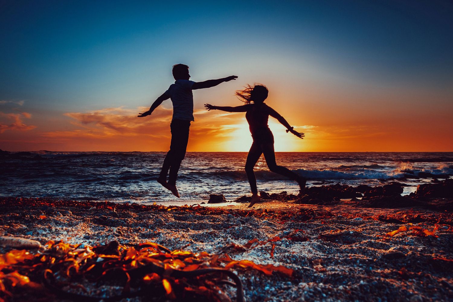 Silhouette of a couple jumping together on a rocky beach during a stunning colorful sunset.