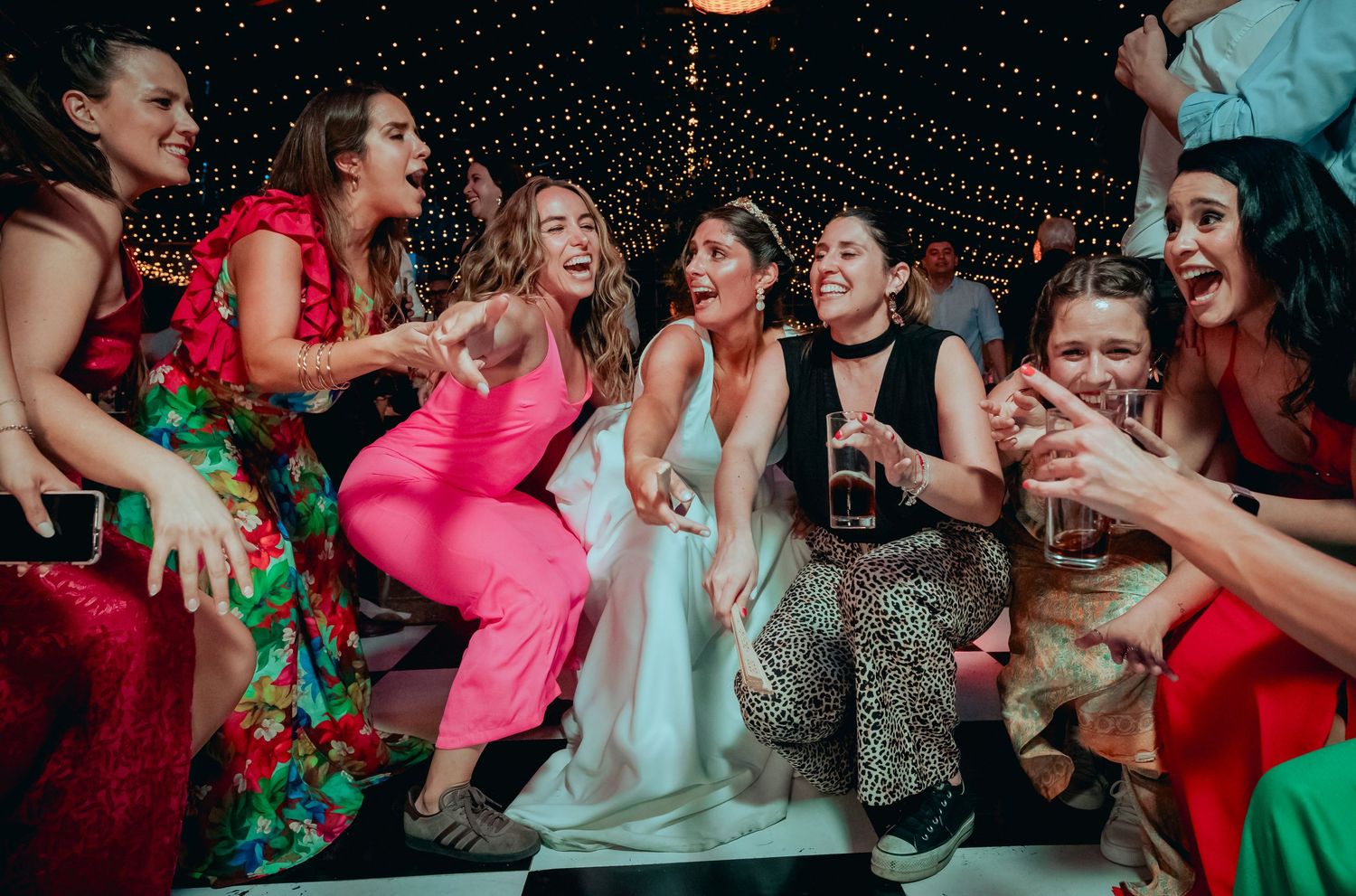 Bride laughs joyfully with group of female friends in colorful outfits at wedding reception party.