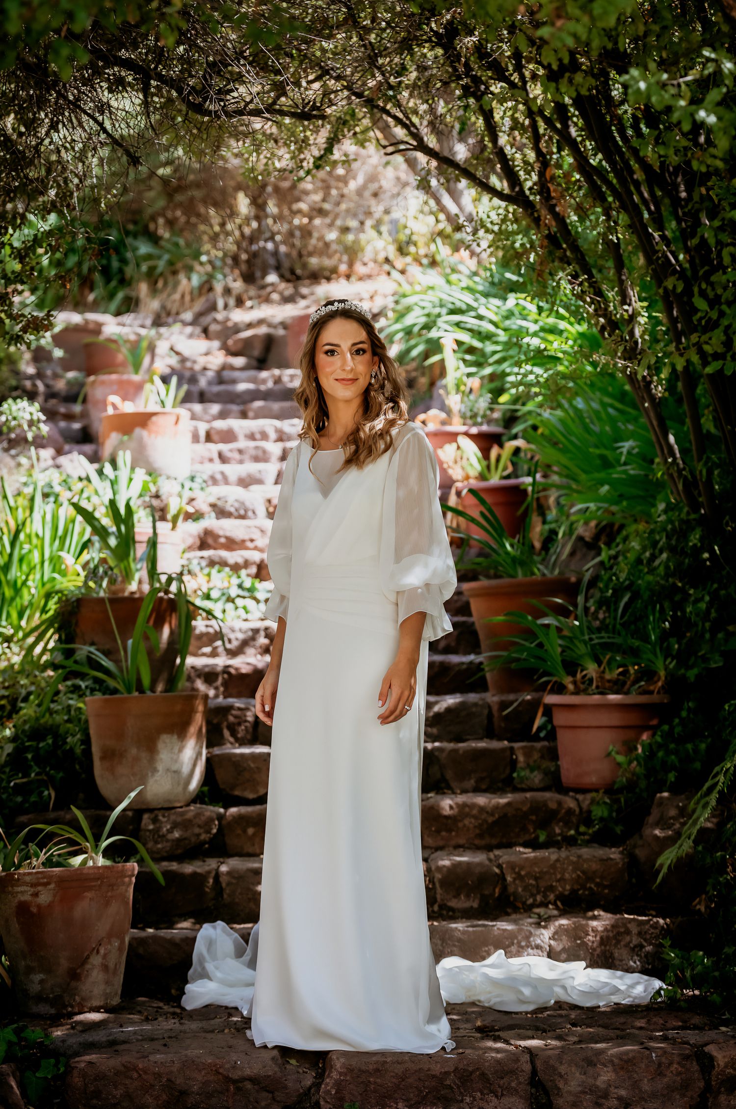 Bride in a simple white wedding dress with sheer cape stands among lush greenery and stone steps.