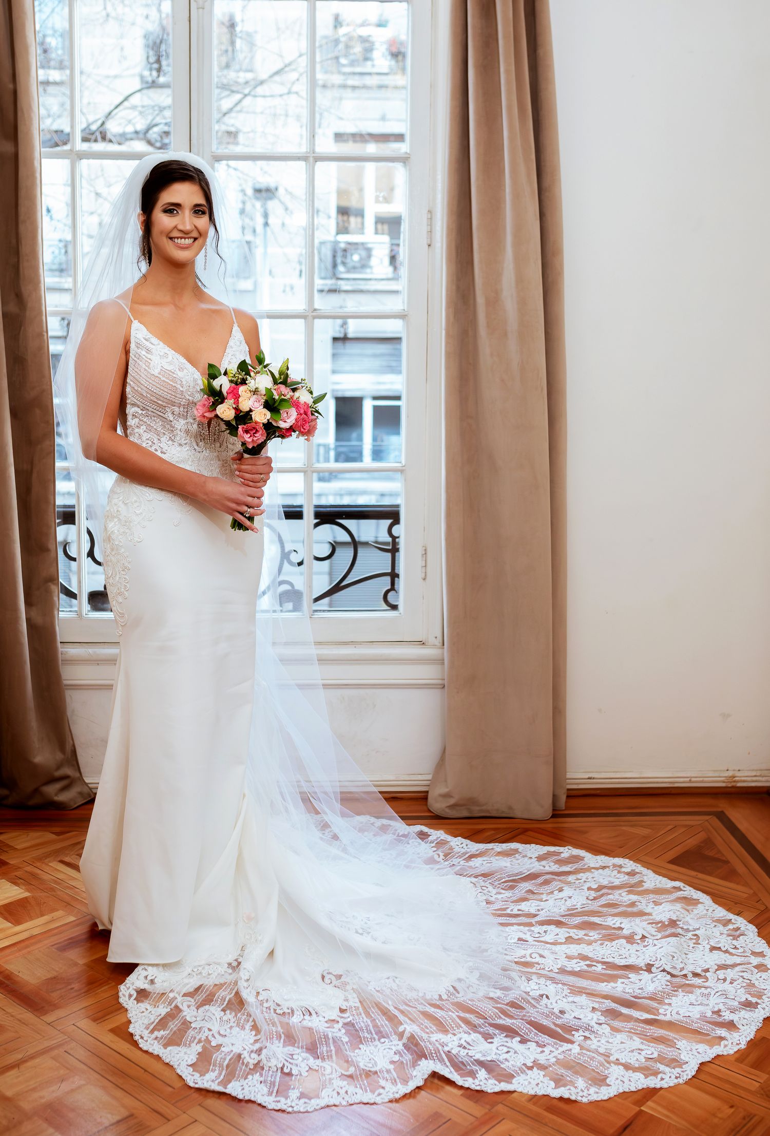 Bride in white lace train gown holding pink bouquet stands before window in elegant room.