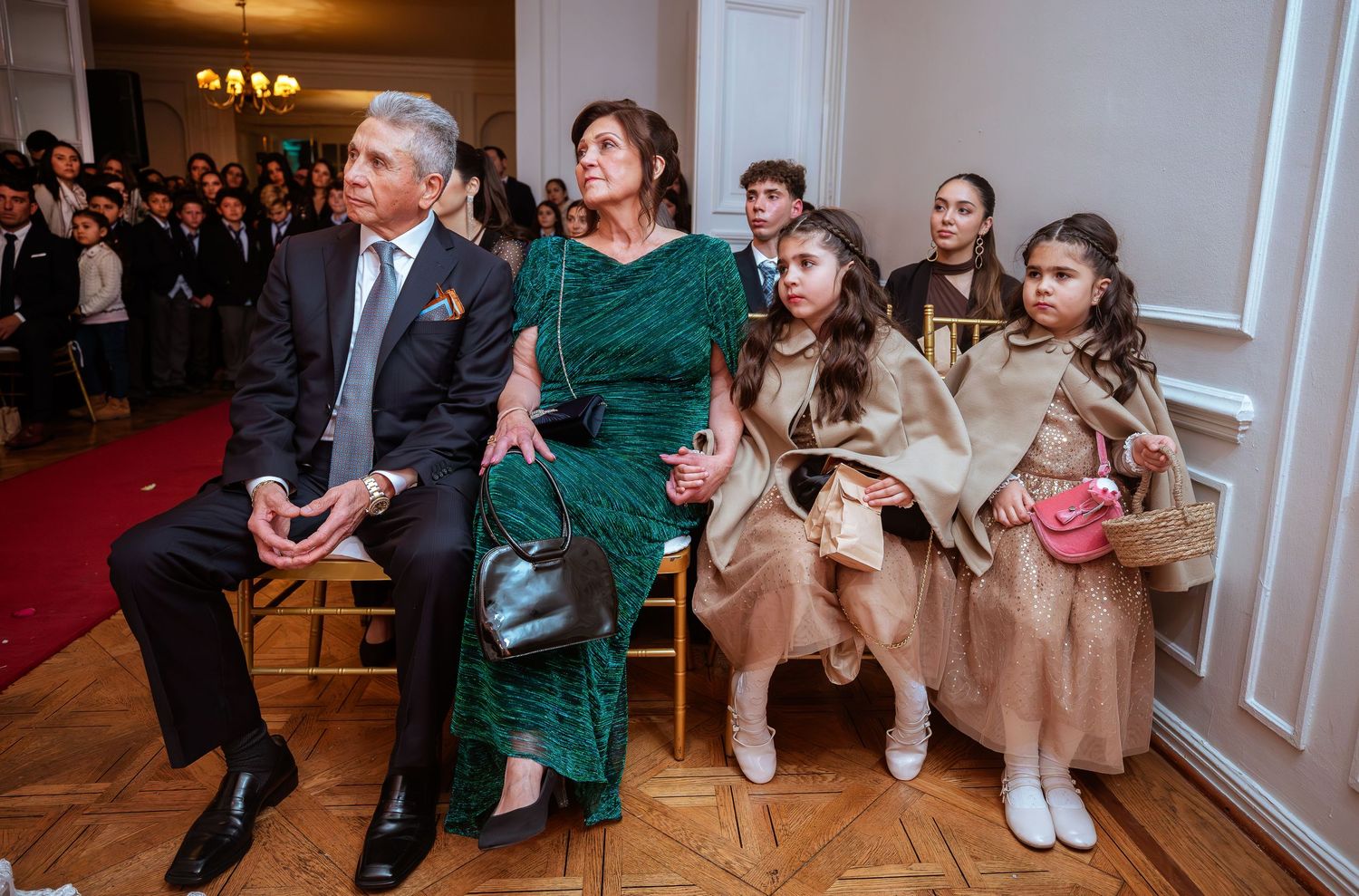 Elegantly dressed family at formal event, grandparents seated with young girls in gold dresses holding pink purses.