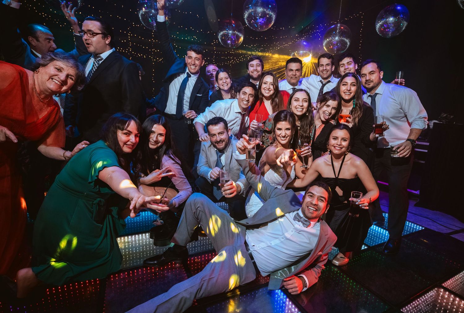 A large group of friends celebrating at a nightclub, posing together with drinks under colorful disco lights.