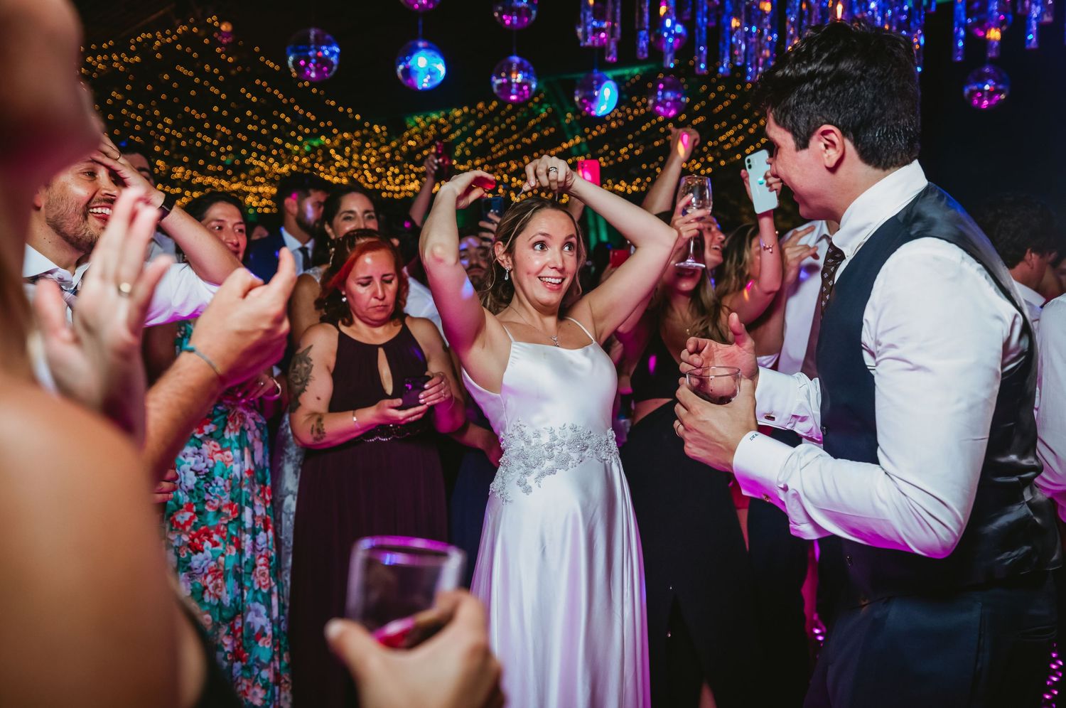 A radiant bride in a white gown dances and smiles with guests on a colorful dance floor at her wedding reception.