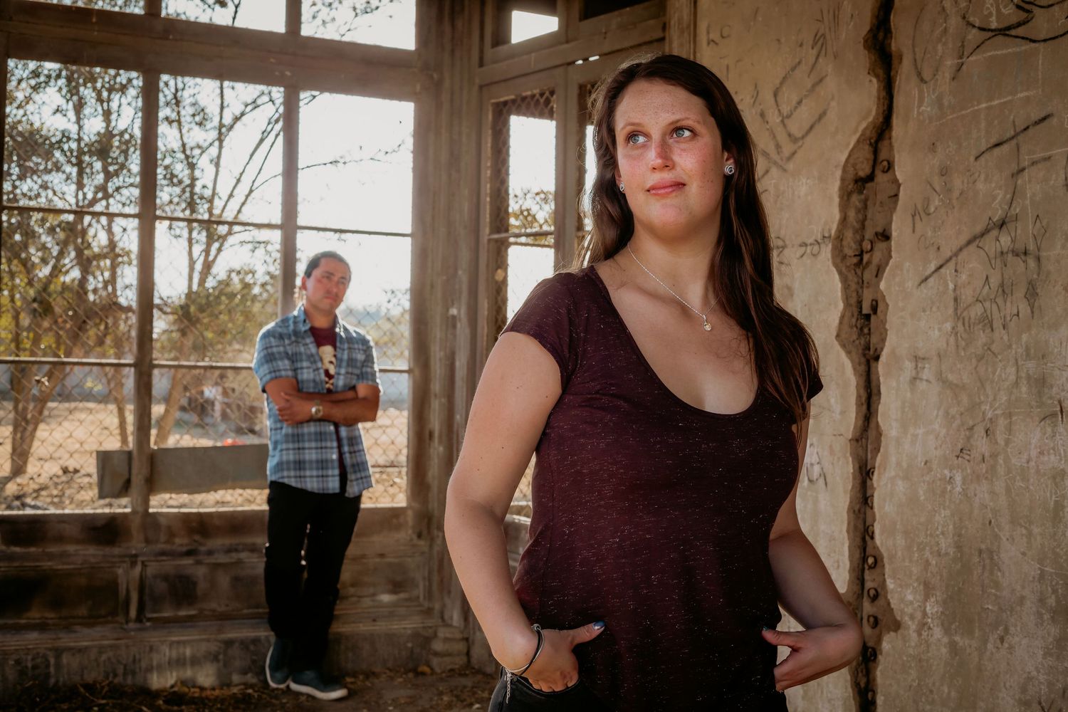 Woman in maroon top poses in abandoned building with man standing by sunlit window behind her.