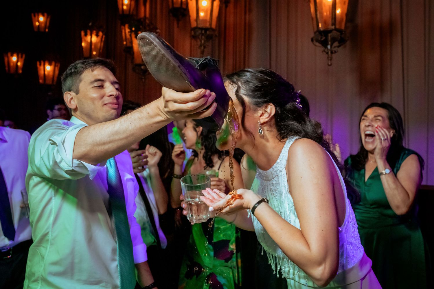 Wedding guest pours drink from a shoe over a laughing woman while guests react with amusement.