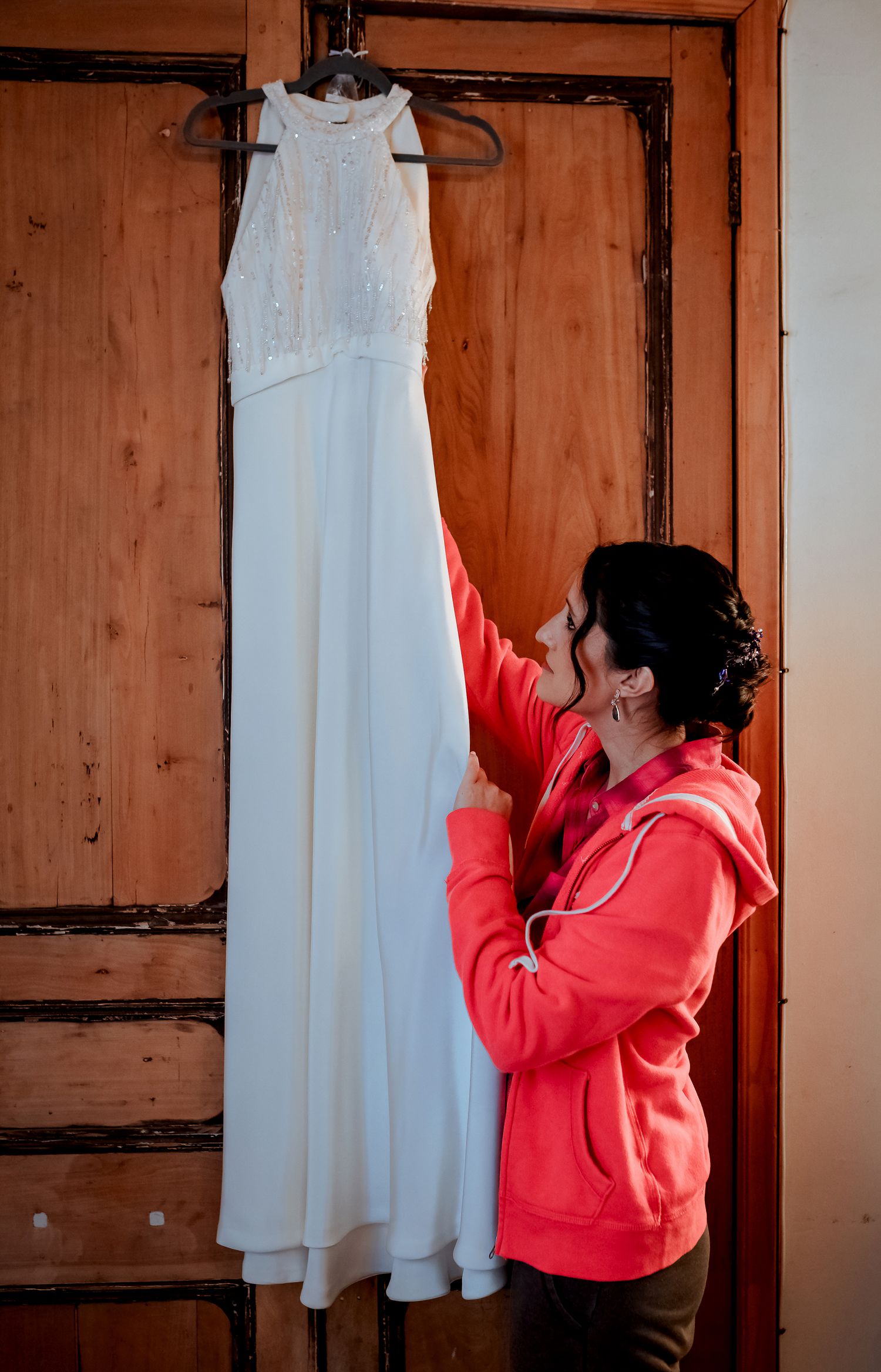 Bride in pink hoodie admiring white halter wedding dress hanging on wooden door.