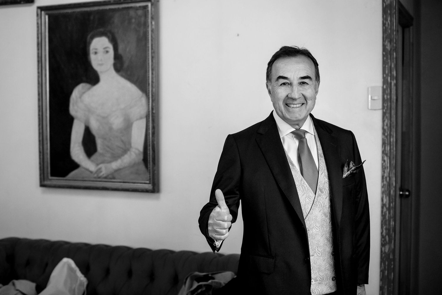 Smiling man in suit giving thumbs up beside classic portrait painting in black and white.